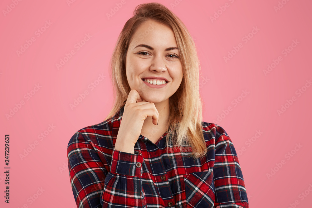 Portrait of good looking blonde female with gentle smile, keeps one hand under chin, dressed in checkered shirt, isolated over pink background. Photo of attractive woman models in studio alone Portrait of good looking blonde female with gentle smile, keeps one hand under chin, dressed in checkered shirt, isolated over pink background. Photo of attractive woman models in studio alone
