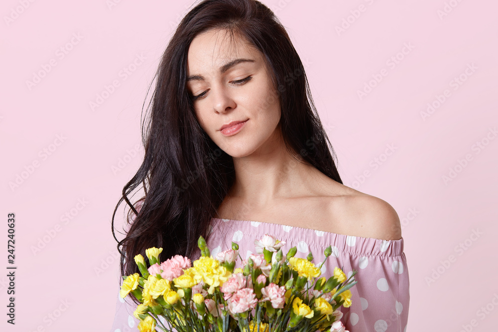 Cropped shot of pleasant looking young woman with dark hair, dressed in polka dot dress, holds bouquet of yellow, pink and white flowers, isolated over pink studio background. Bouquet for 8 March Cropped shot of pleasant looking young woman with dark hair, dressed in polka dot dress, holds bouquet of yellow, pink and white flowers, isolated over pink studio background. Bouquet for 8 March
