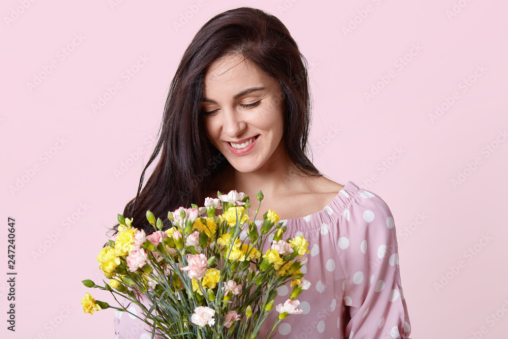 Positive of young European woman with dark hair, holds pretty bouquet, smiles gently, wears polka dot fashionable clothes, prepares for International Womens Day, isolated over pink background