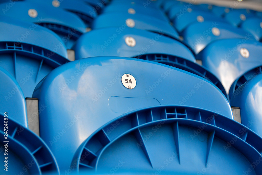 Obraz premium Blue plastic stadium seats with silver badges marking the seat numbers. Empty seats with focus on number 54.