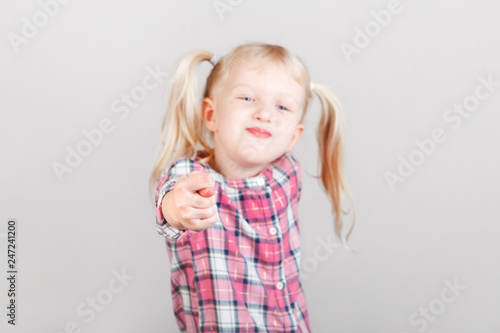 Cute adorable white blonde Caucasian preschool girl making faces in front of camera. Child showing fig sign and posing in studio on plain light background. Kid expressing negative emotions