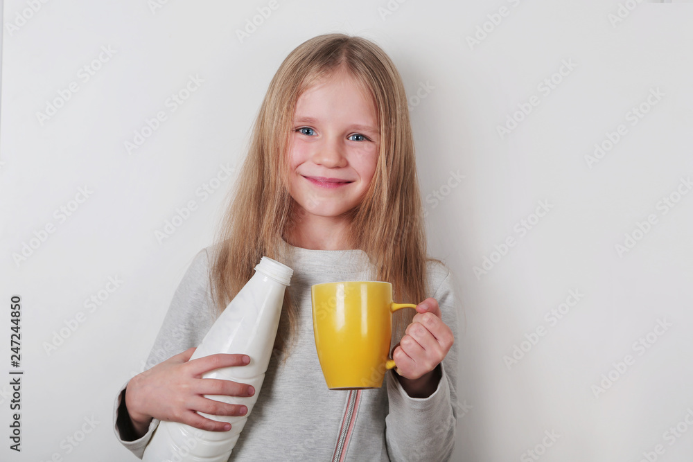 Cute happy blond little girl drinking milk or yogurt from a yellow cup on a white background.  Girl holding cup and plastic bottle. Dairy products concept