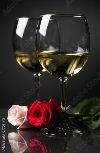 Two glasses of white wine with a pink and red rose on a dark setting.