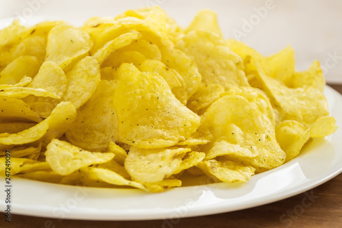 Pile of crispy potato chips in a white plate on a table. Fast food and tasty snack consept. Front view, close up, selective focus