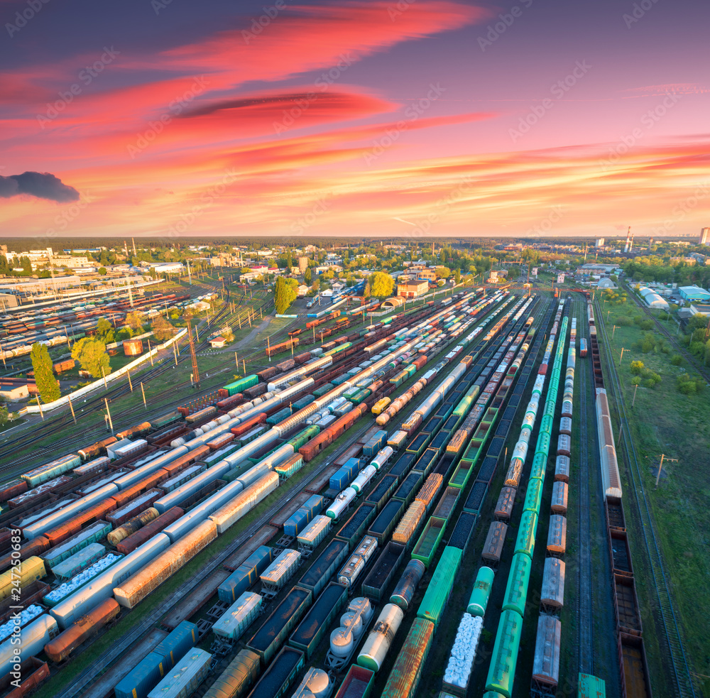 Aerial view of freight trains on railway station at colorful sunset. Wagons with goods on railroad. Heavy industry. Industrial. Cargo trains, city buildings and orange sky with red clouds. Top view - obrazy, fototapety, plakaty 