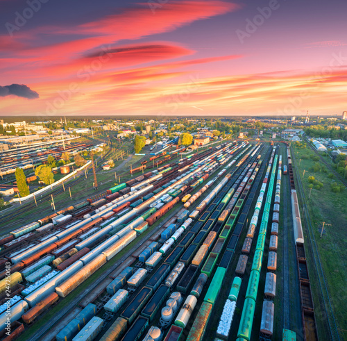 Aerial view of freight trains on railway station at colorful sunset. Wagons with goods on railroad. Heavy industry. Industrial. Cargo trains, city buildings and orange sky with red clouds. Top view photo