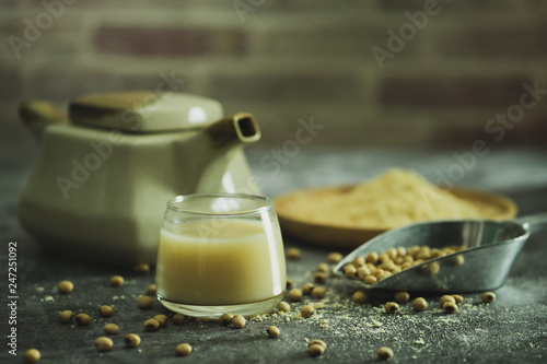 Soymilk in the glass and the kettle is placed beside. Soybean powder is crushed in a wooden dish and has scattered soy beans on the table in morning light.