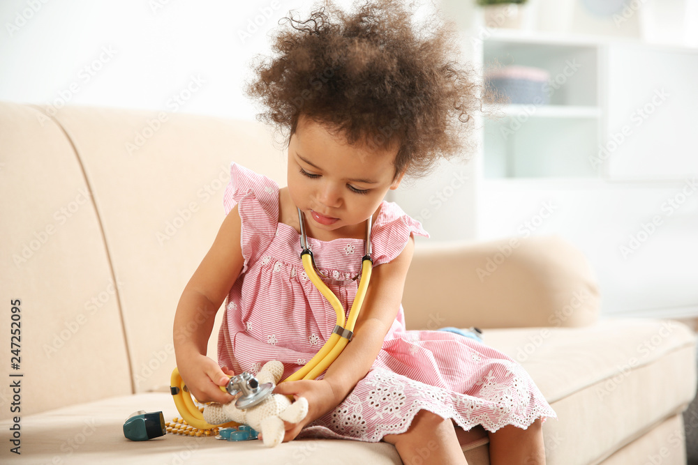Cute African American child imagining herself as doctor while playing with stethoscope and toy on couch at home Cute African American child imagining herself as doctor while playing with stethoscope and toy on couch at home