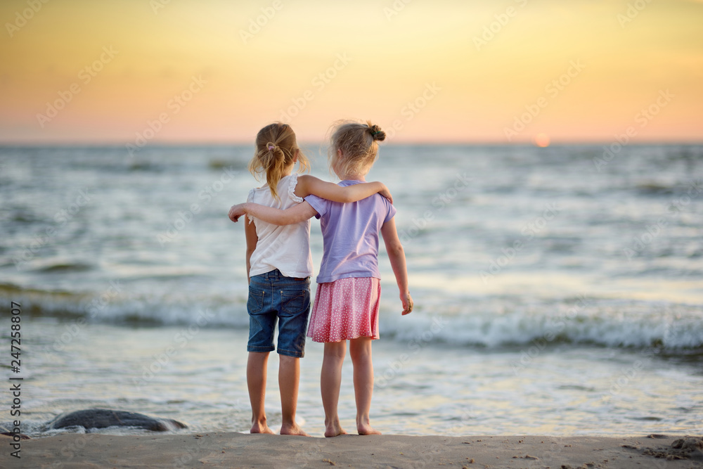 Two cute young sisters having fun on a sandy beach on warm and sunny summer day by the sea. Kids playing by the ocean.