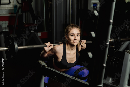 Active beautiful fitness model girl crouches with a barbell on the shoulders in the gym.