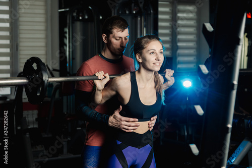 Handsome athletic man helps a beautiful girl to squat with a barbell on the shoulders in the gym.