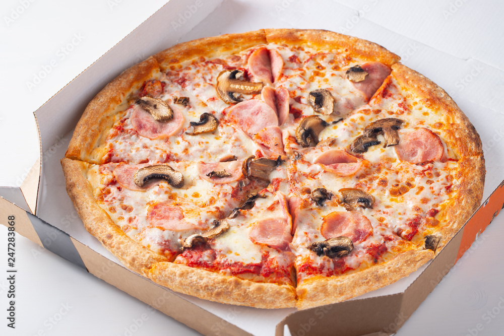 Italian fast food. Delicious hot pizza in a box with ham, champignons sliced and served on white table, close up view. Concept of fast and unhealthy diet