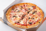Italian fast food. Delicious hot pizza in a box with ham, champignons sliced and served on white table, close up view. Concept of fast and unhealthy diet