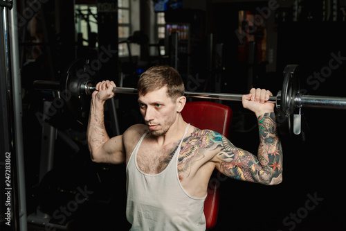 Handsome athletic man trains shoulders lifting barbell sitting on the bench in the gym.