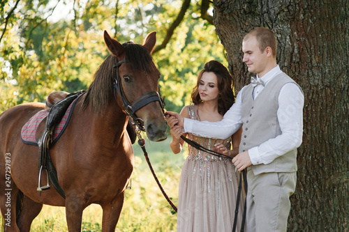 Wedding at sunset. Couple is sitting on a bench under a tree. Beige dress with sparkles. Light suit with a bow tie. The bride and groom embrace and kiss.