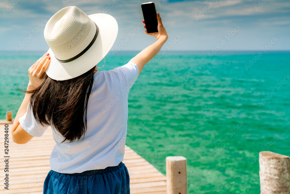 Young Asian woman wear straw hat in casual style use smartphone taking selfie at wooden pier. Summer vacation at tropical paradise beach. Happy girl travel on holiday. Woman enjoy and relax life.