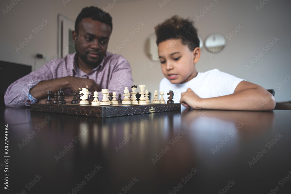 Cute african american son playing chess with his black father. The son teaches his father to play chess. Father teaches his son to play chess. Hobby for children. Cute african american son playing chess with his black father. The son teaches his father to play chess. Father teaches his son to play chess. Hobby for children.