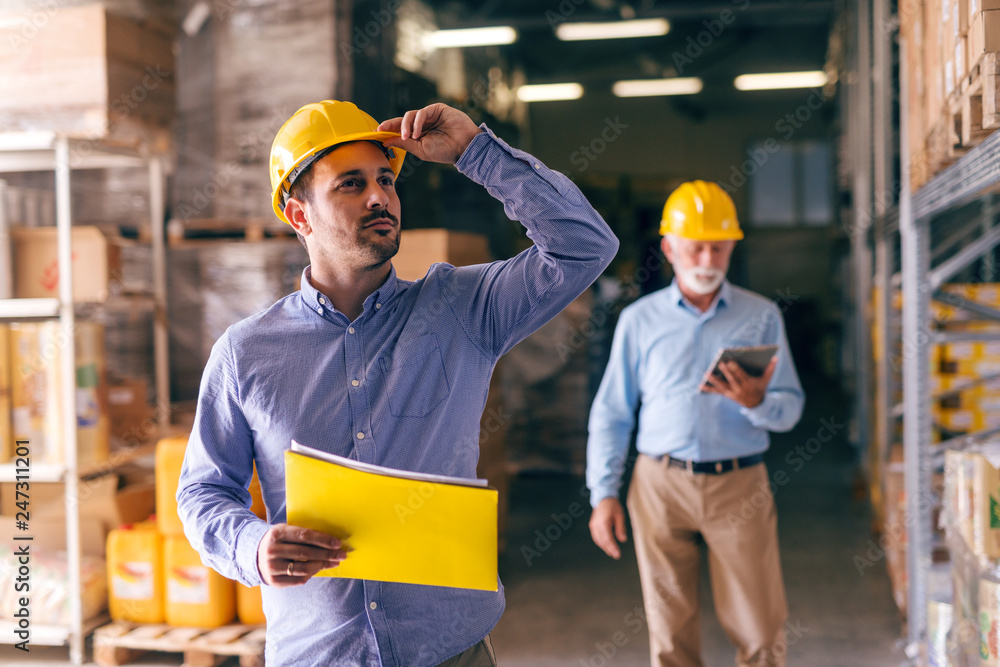 Businessman holding helmet and folder with documents while looking up and standing in warehouse. In background his colleague walking with tablet. Businessman holding helmet and folder with documents while looking up and standing in warehouse. In background his colleague walking with tablet.