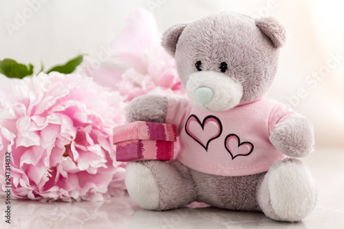 Little grey teddy bear sitting with a gift box and pink peonies behind him. Romantic gift ideas, presents for children.