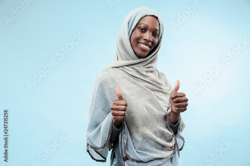 The beautiful young black african muslim girl wearing gray hijab at blue studio. She standing with a happy smile on her face. The human emotions, facial expression concept. Trendy colors photo