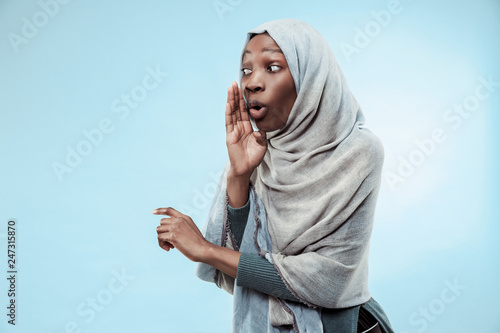Secret, gossip concept. Young african woman whispering a secret behind her hand. The woman isolated on trendy blue studio background. Young emotional woman. Human emotions, facial expression concept. photo
