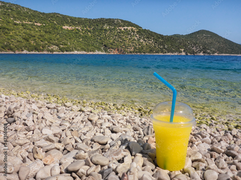 A glass of refreshing cold summer citrus lemon drink stands on white stones at pebble beach on the background of turquoise sea Antisamos beach, Kefalonia island, Greece. A glass of refreshing cold summer citrus lemon drink stands on white stones at pebble beach on the background of turquoise sea Antisamos beach, Kefalonia island, Greece.