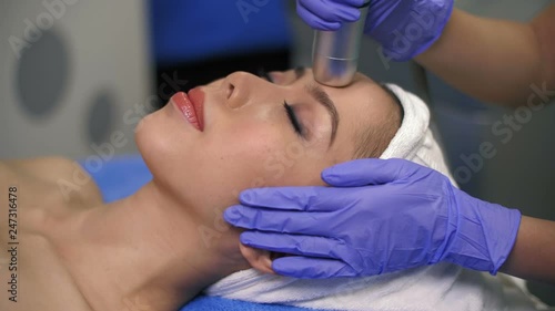 Close up of Asian woman with closed eyes lying on treatment table while beautician massaging her face with special equipment photo