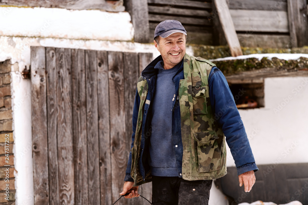 Smiling senior man dressed in warm clothes holding bucket and looking away. Countryside exterior.