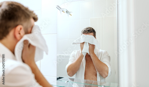 Handsome businessman with unbuttoned shirt wiping his face with towel while standing inn bathroom in front of mirror. Morning routine concept.