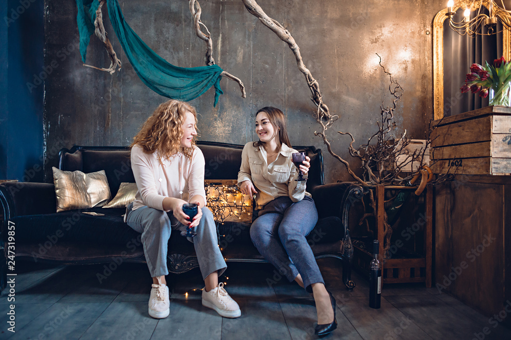 Two friends or sisters enthusiastically discuss something sitting on the sofa in a cozy festive atmosphere over a glass of wine.