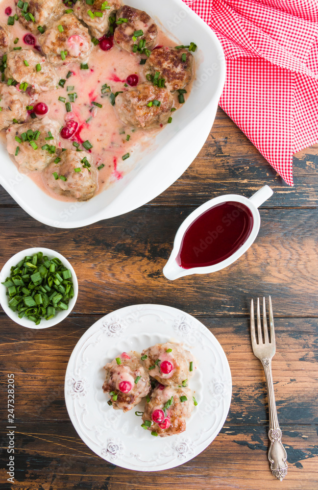 Swedish meatballs in a red cowberry sauce with green onion. White casserole and plate on wooden rustic table, top view
