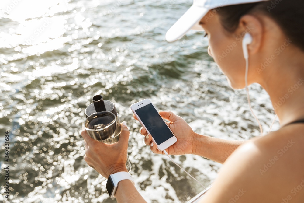 Beautiful young sports fitness woman using mobile phone drinking water at the beach outdoors listening music with earphones.