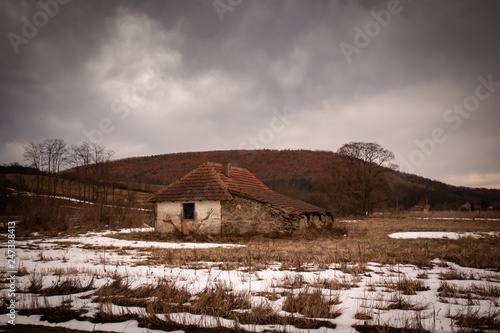 Old small abandoned house on meadow.Winter rural scene at sunset with small bits of snow.