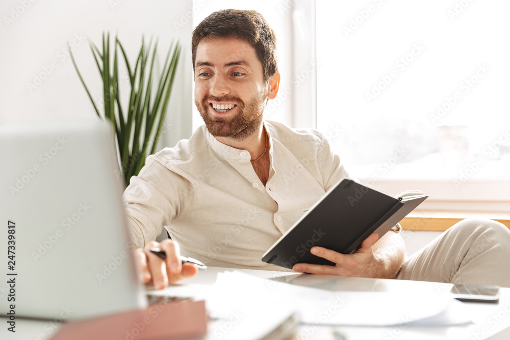 Image of european office worker 30s wearing white shirt using laptop and notebook, while sitting at table in modern workplace Image of european office worker 30s wearing white shirt using laptop and notebook, while sitting at table in modern workplace