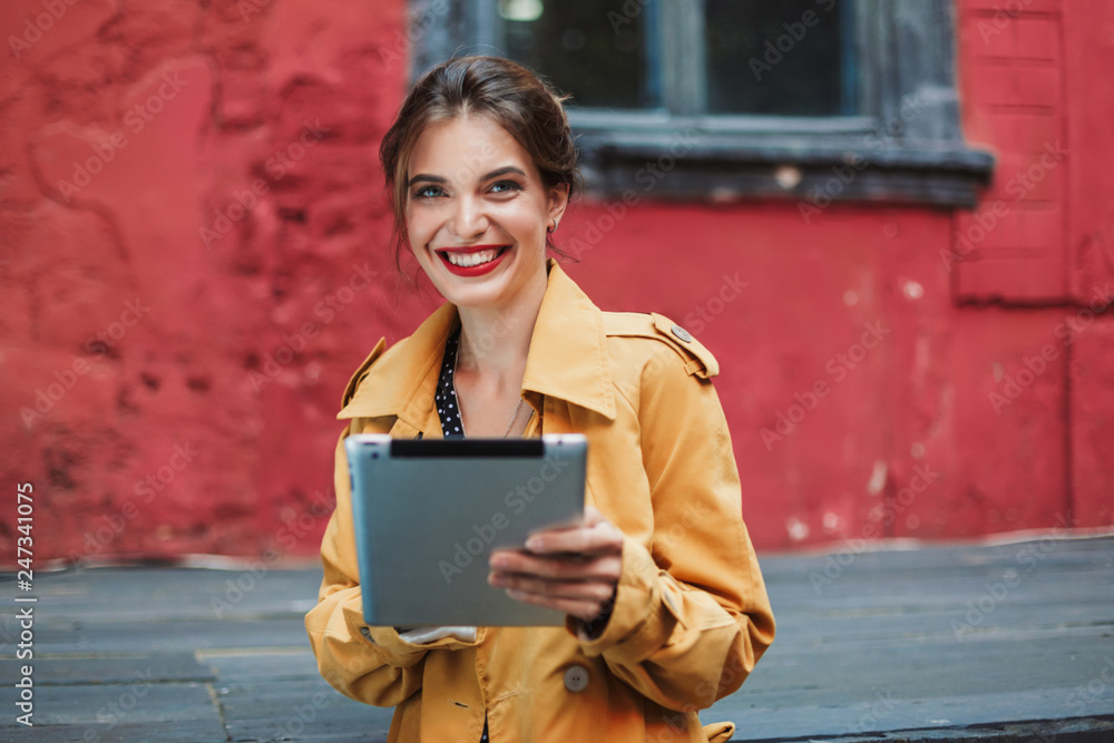 Young attractive cheerful woman in orange trench coat and black polka dot dress holding the tablet in hands while happily looking in camera in old courtyard Young attractive cheerful woman in orange trench coat and black polka dot dress holding the tablet in hands while happily looking in camera in old courtyard