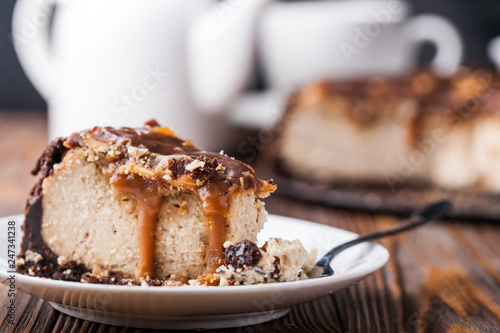 Close up view of a piece of peanut butter chocolate caramel cheesecake pie and cup of coffee on wooden background with a coffee pot and cheesecake in the background. A Sweet Coffee Break concept 