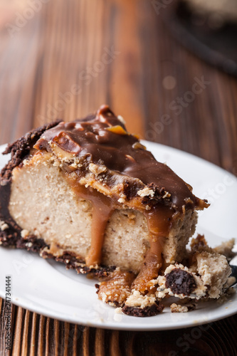 Close up view of a piece of peanut butter chocolate caramel cheesecake pie and cup of coffee on wooden background with a coffee pot and cheesecake in the background. A Sweet Coffee Break concept 