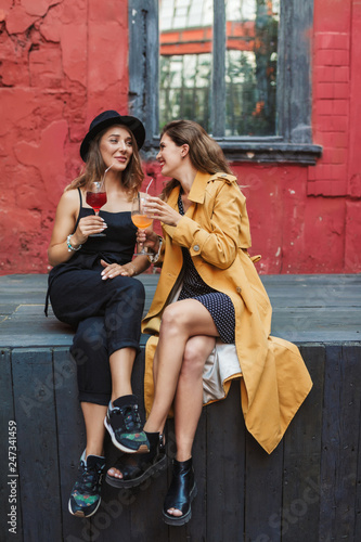 Two young attractive joyful women with cocktails in hands happily talking while spending time together in old cozy courtyard of cafe
