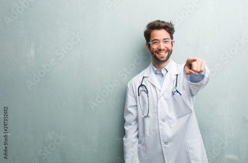 Young friendly doctor man against a grunge wall with a copy space cheerful and smiling pointing to the front