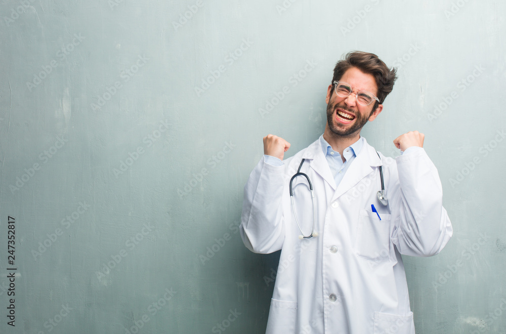 Young friendly doctor man against a grunge wall with a copy space very happy and excited, raising arms, celebrating a victory or success, winning the lottery Young friendly doctor man against a grunge wall with a copy space very happy and excited, raising arms, celebrating a victory or success, winning the lottery
