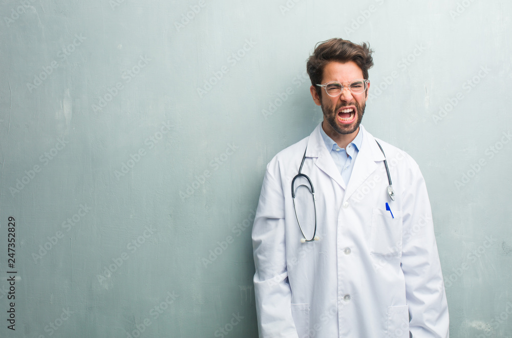 Young friendly doctor man against a grunge wall with a copy space screaming angry, expression of madness and mental instability, open mouth and half-opened eyes, madness concept Young friendly doctor man against a grunge wall with a copy space screaming angry, expression of madness and mental instability, open mouth and half-opened eyes, madness concept