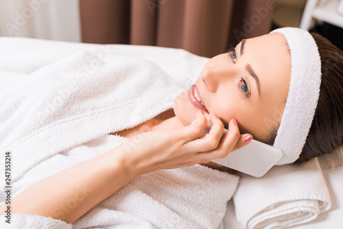 attractive woman lying in white bathrobe and talking on smartphone at beauty salon