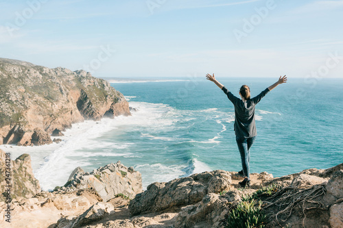 The girl next to the Atlantic Ocean in Portugal raises up her hands shows how pleased she is.