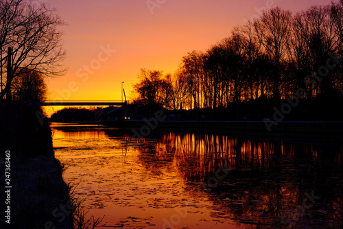Dramatic and colorful sunrise over a Beautiful early winter landscape with a frozen river or canal, treelined riverside and grass at sunrise creating a tranquile and quiet scenic nature background