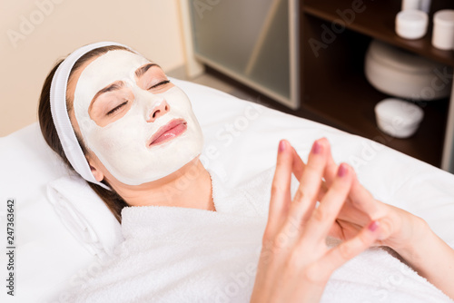 woman lying in white bathrobe and hairband with applied facial mask at beauty salon
