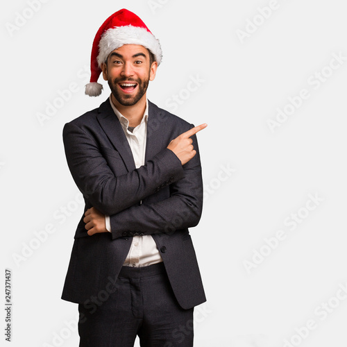 Young business man wearing santa hat pointing to the side with finger © Asier