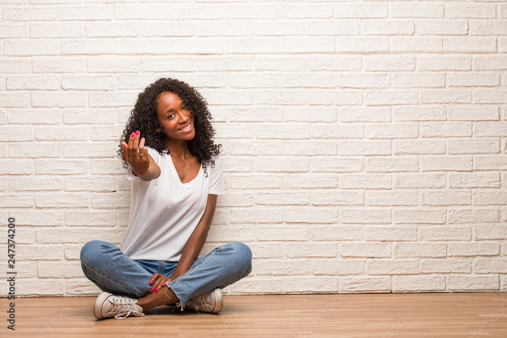 Young black woman sitting on a wooden floor inviting to come, confident and smiling making a gesture with hand, being positive and friendly Young black woman sitting on a wooden floor inviting to come, confident and smiling making a gesture with hand, being positive and friendly