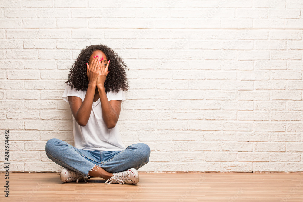 Young black woman sitting on a wooden floor feels worried and scared, looking and covering face, concept of fear and anxiety Young black woman sitting on a wooden floor feels worried and scared, looking and covering face, concept of fear and anxiety