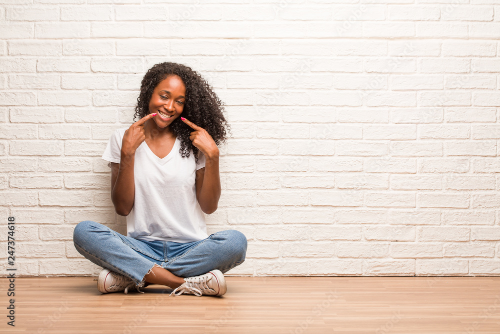 Young black woman sitting on a wooden floor smiles, pointing mouth, concept of perfect teeth, white teeth, has a cheerful and jovial attitude Young black woman sitting on a wooden floor smiles, pointing mouth, concept of perfect teeth, white teeth, has a cheerful and jovial attitude