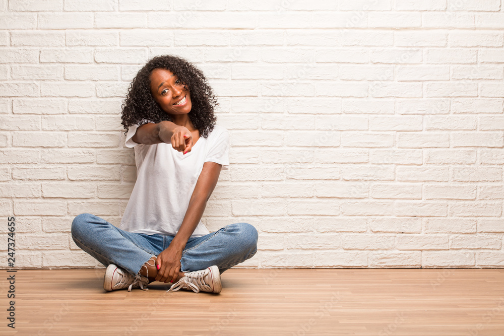 Young black woman sitting on a wooden floor cheerful and smiling pointing to the front Young black woman sitting on a wooden floor cheerful and smiling pointing to the front
